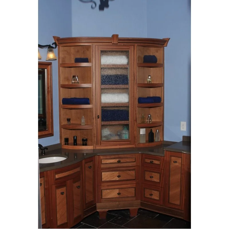 A wooden bathroom cabinet with open shelves, some with folded towels and decorative items, a glass-paneled central door, and a countertop with cabinets below, set against a light blue wall.
