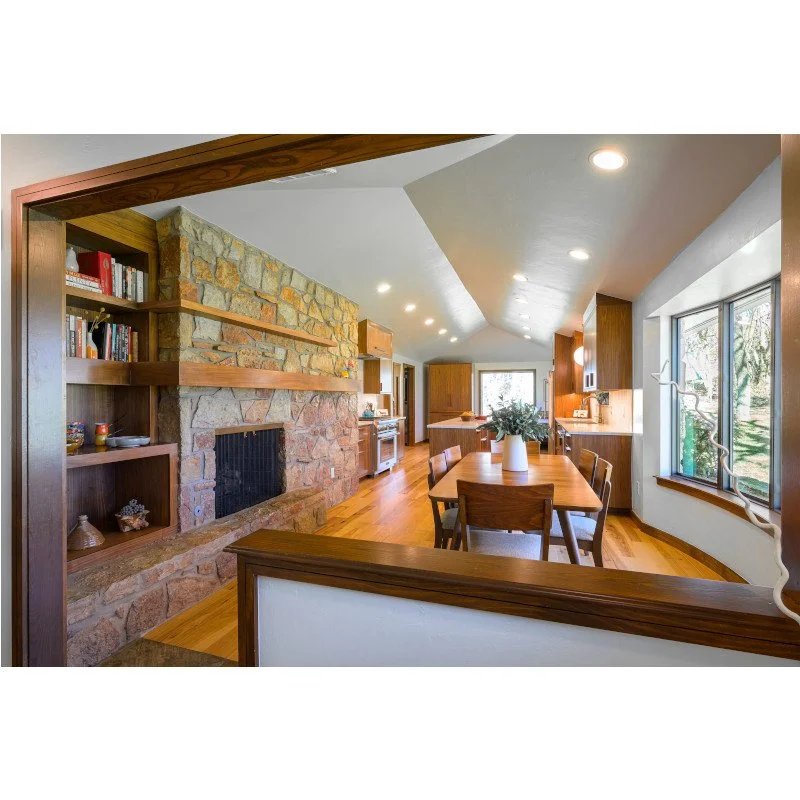 View of a kitchen and dining area with a stone fireplace, wooden shelving, a dining table with chairs, and large windows with a garden view.