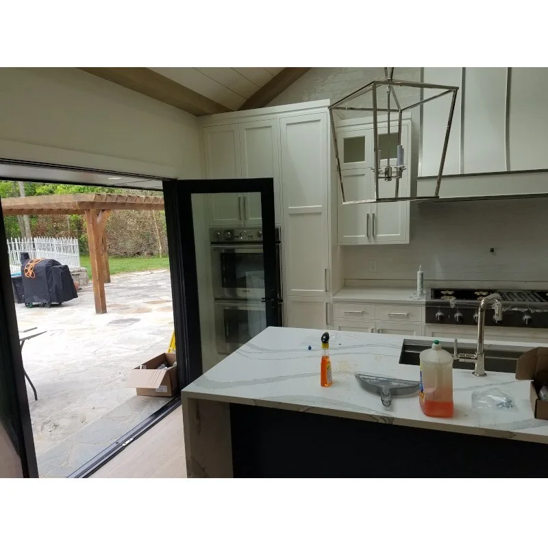 Modern kitchen with white cabinets, marble countertop island, built-in double oven, and stainless steel stove, overlooking backyard patio through sliding glass door.