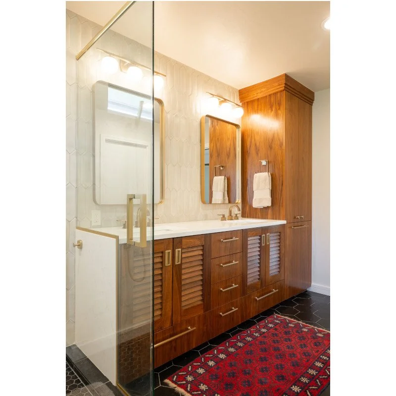 Bathroom with double vanity, wooden cabinets, two mirrors, towel hooks, and a red patterned rug on black hexagonal tile floor.
