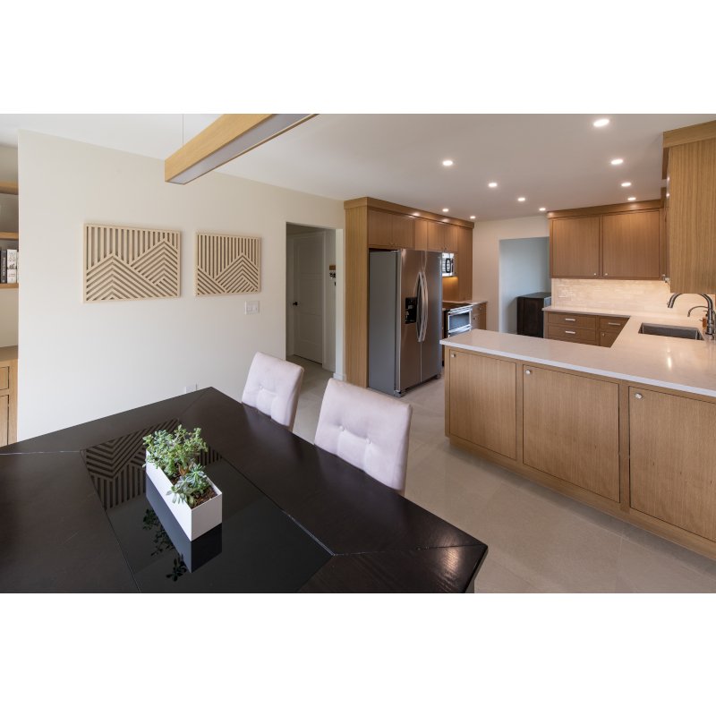 Modern kitchen with wooden cabinets, stainless steel refrigerator, and breakfast bar, adjacent to a dining table with chairs and a decorative plant.