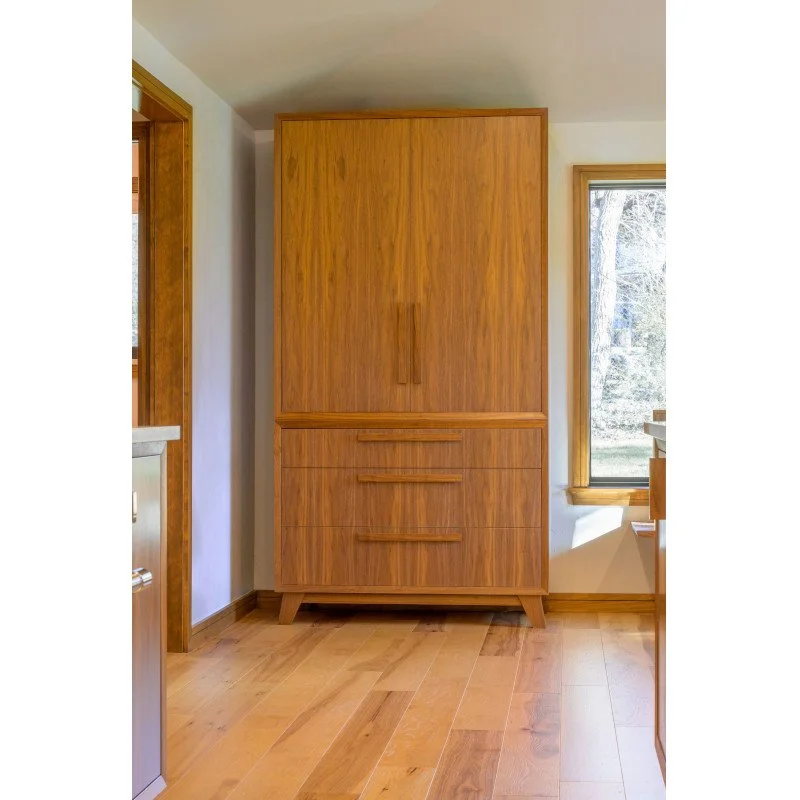 A wooden cabinet with a mid-century modern design, featuring three drawers at the bottom and two large doors at the top, situated in a room with wooden flooring and a window to the right.