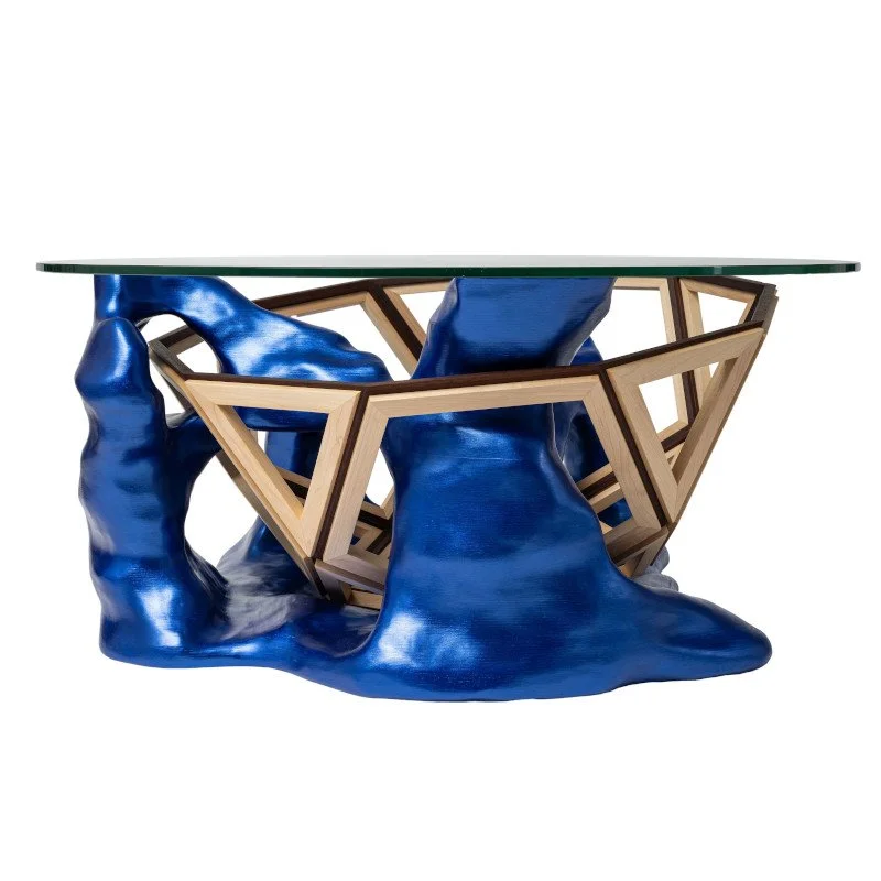 Modern glass-top table with a sculptural base made of blue, rock-like forms and wooden geometric accents.