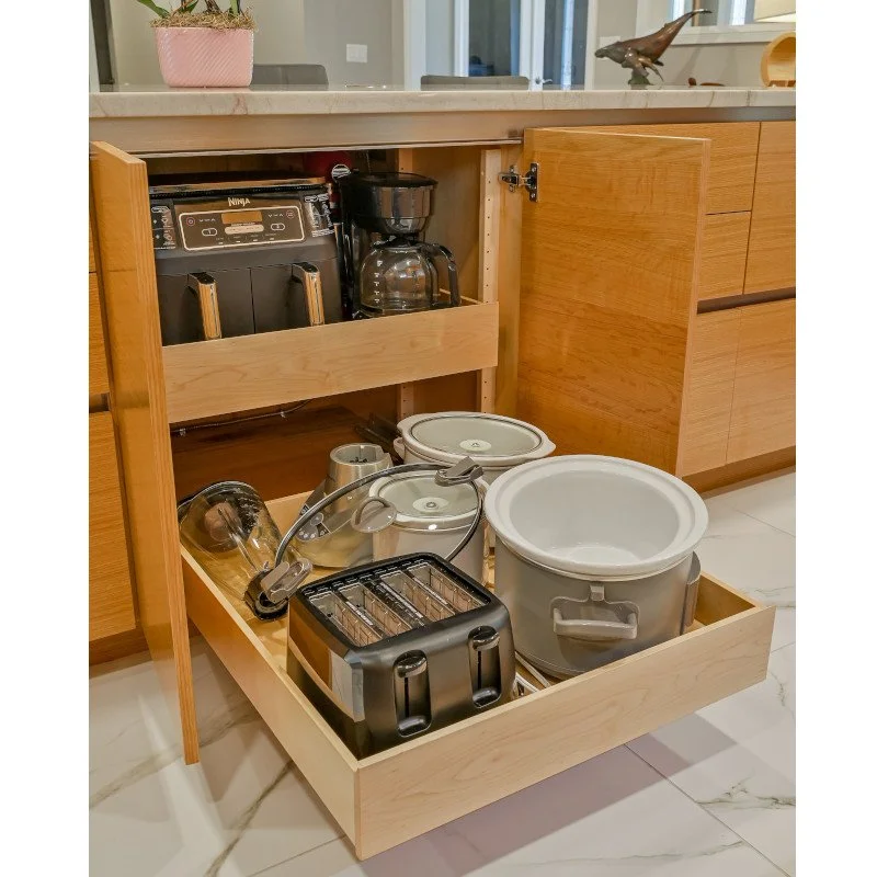 Open kitchen cabinet drawer containing kitchen appliances such as a toaster, slow cooker, and coffee maker, with a countertop and decorative items in the background.
