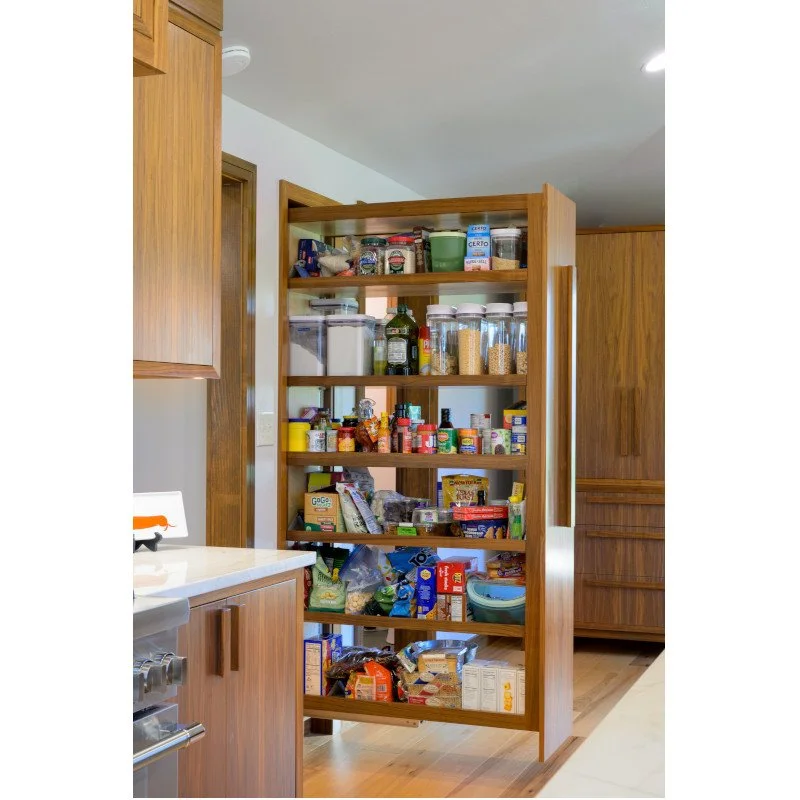 A wooden pull out pantry with multiple shelves containing canned goods, jars, bottles, boxes, and various grocery items in a kitchen.