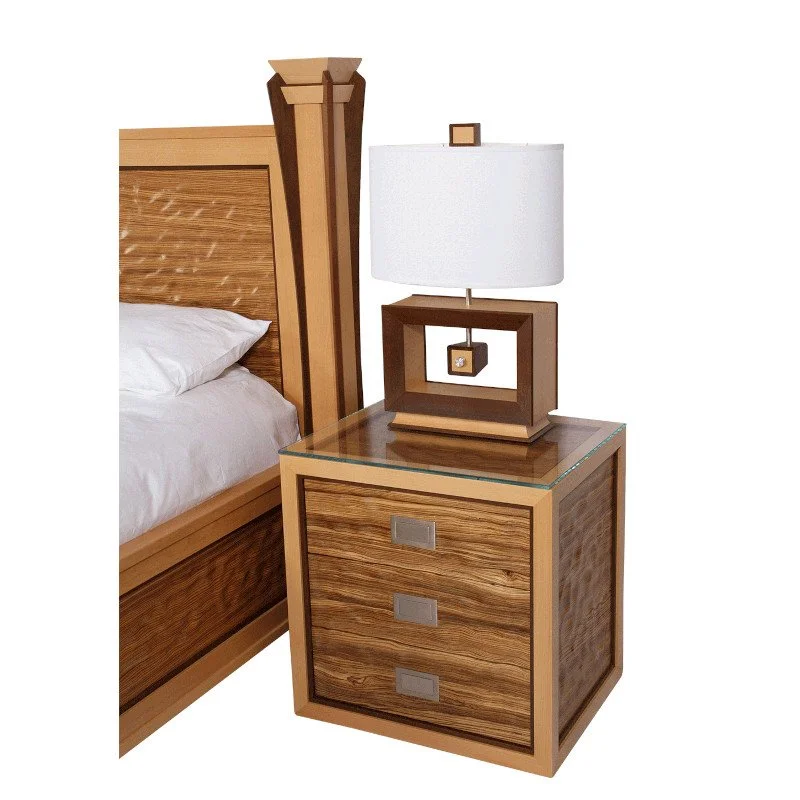 A wooden nightstand with three drawers, a modern lamp with a white shade and wooden base, and a bed with a wooden headboard.