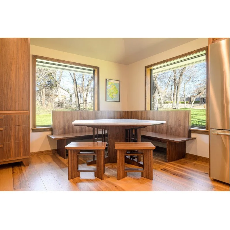 Dining nook with a corner bench, table, and two stools in a kitchen with large windows showing an outdoor yard with trees.