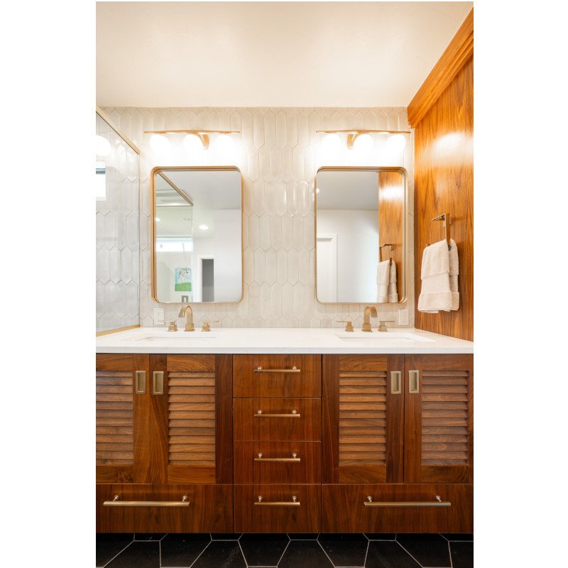 Bathroom vanity with two mirrors, wooden cabinets, and white countertops with sinks, framed by wall-mounted lights, and a wooden wall on the right with towel hooks holding white towels.