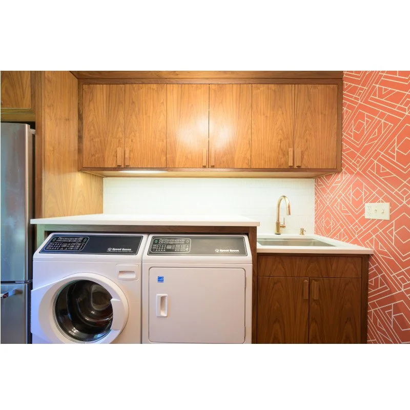 Laundry room with a front-loading washing machine, a dryer, wooden cabinets, a small sink with a gold faucet, and a decorative pink wall with geometric patterns.