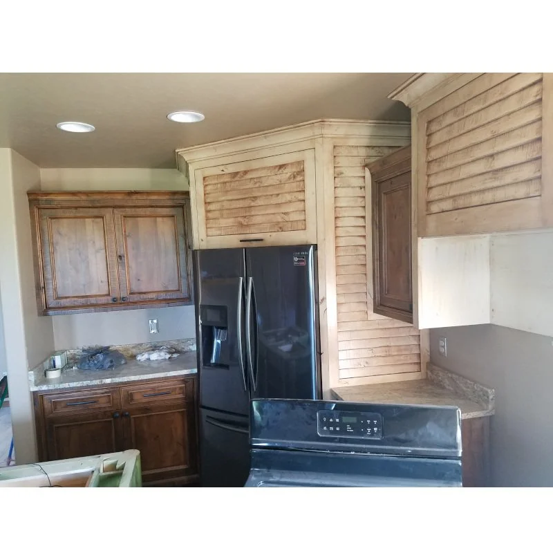 Rustic kitchen with wooden cabinets, stainless steel refrigerator, and a black stove, with beige walls and ceiling lights.