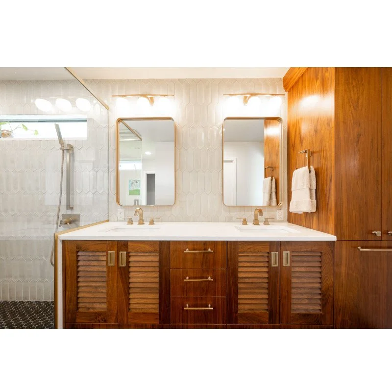 A bathroom with two mirrors above a double sink vanity with wooden cabinets, a towel hanging on the wall, and a walk-in shower with a handheld showerhead.