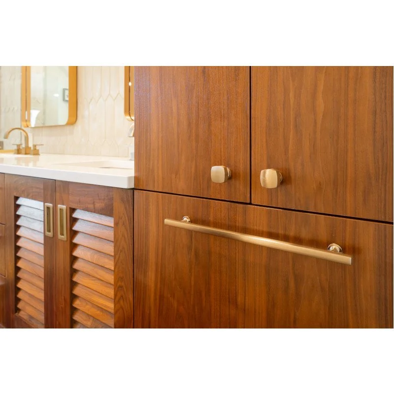 Close-up of wooden bathroom cabinets with louvered doors, a gold handle, and a gold knob, with a section of white countertop and a mirror in the background.