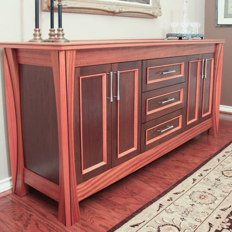A wooden sideboard with dark brown doors and drawers, framed with lighter wood trim, located on a hardwood floor next to a beige and red patterned rug.