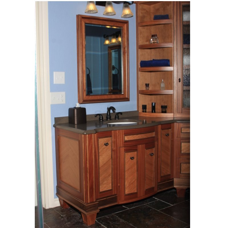 Wooden bathroom vanity with a gray countertop, a mirror above, a dark soap dispenser, and a towel and small containers on open shelving next to the mirror.