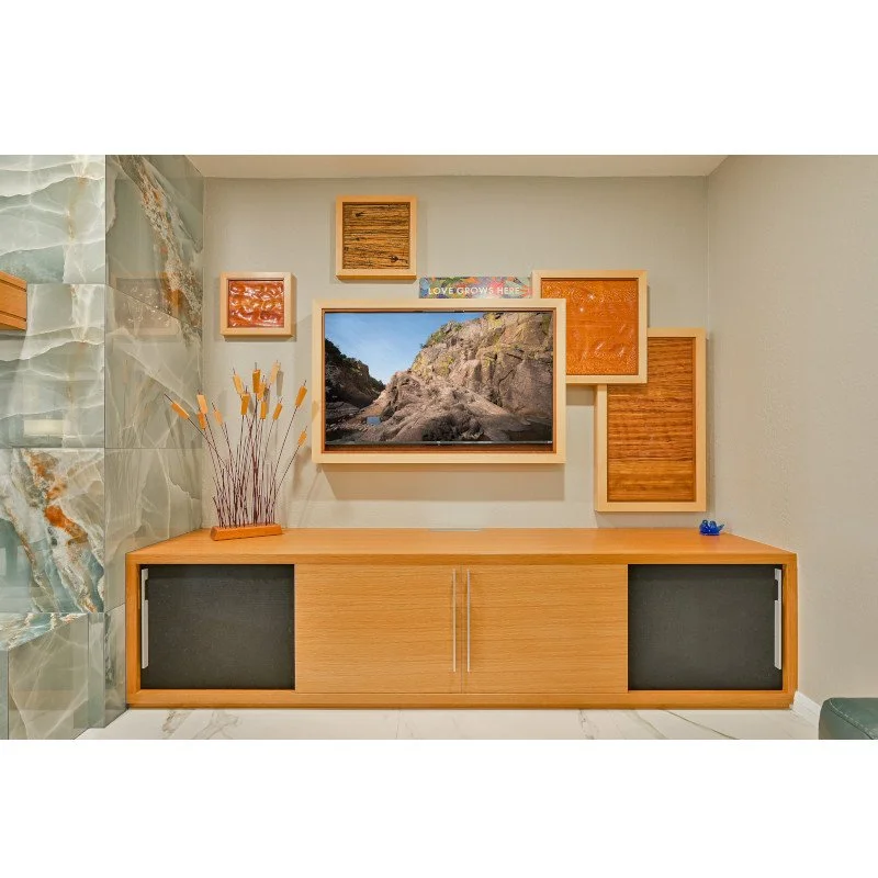 Living room entertainment center with a wall-mounted TV surrounded by framed wooden wall art, a wood art piece resembling cattails or reeds, and a small blue figurine, against a wall of marble tiles and plain painted wall.