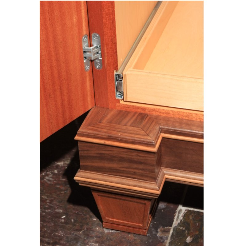 Close-up of a corner of a wooden cabinet with a decorative step at the bottom, showing a metal hinge attached to the door.