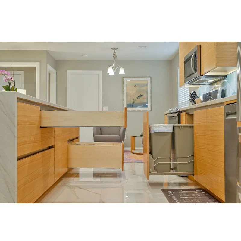Kitchen with open cabinets, a gray armchair in the background, and a wall art with birds, featuring light wood cabinets, a microwave, and a garbage bin.