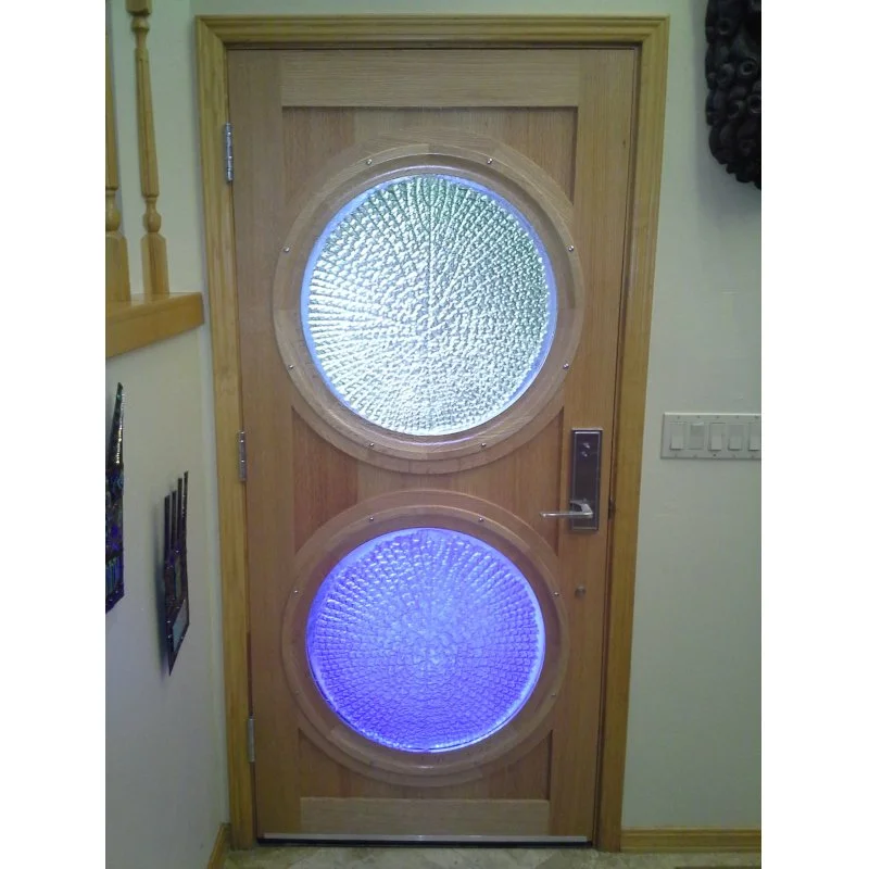 A wooden door with two circular art glass windows, one above the other, back lit from sun light, in an indoor setting with a light switch on the right side.