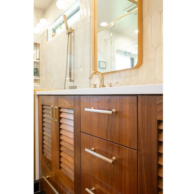 Bathroom vanity with wooden cabinet, gold accents, and a white countertop with a gold faucet. A mirror above and a window with natural light. Part of the shower area with a squeegee and shelves visible.