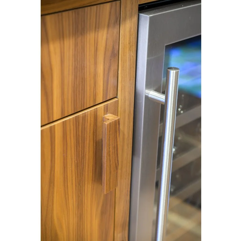 Close-up of a wooden kitchen cabinet door with a vertical, rectangular handle next to a stainless steel wine fridge with a long, rounded handle.