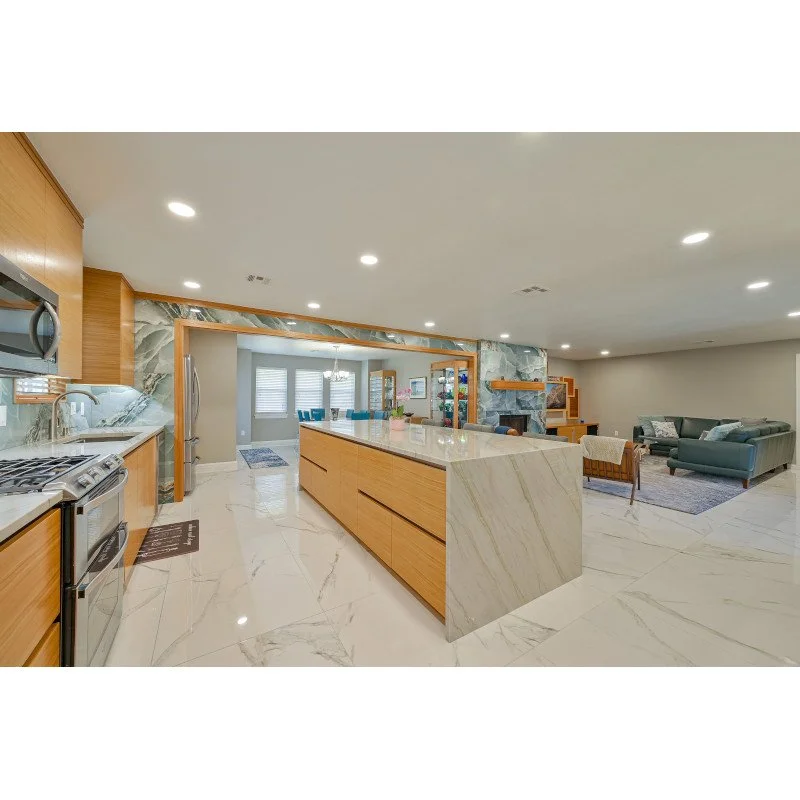 Open-concept kitchen with quartzite countertops, wooden cabinets, stainless steel appliances, and a large island, leading to a living room with sofas and an entertainment center in the background.