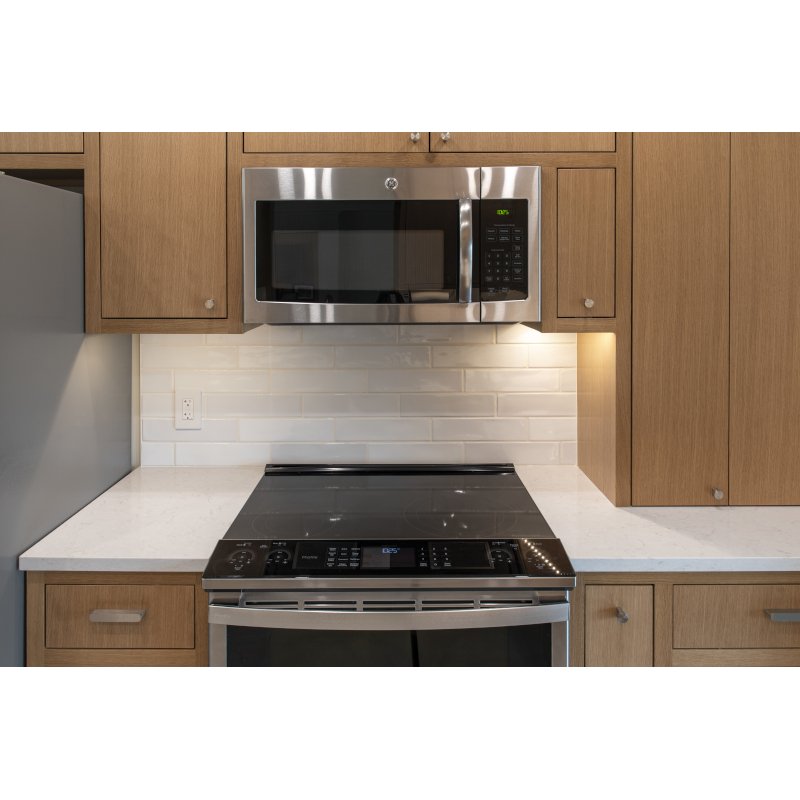 Kitchen with wooden cabinets, a stainless steel microwave above the stove, a white countertop, and a white tiled backsplash.