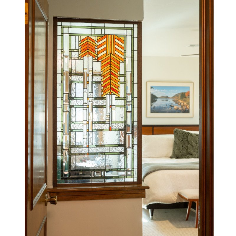 A stained glass window with geometric patterns and orange arrow-shaped details, located next to a bedroom with a bed, a framed landscape picture, and a green textured pillow.