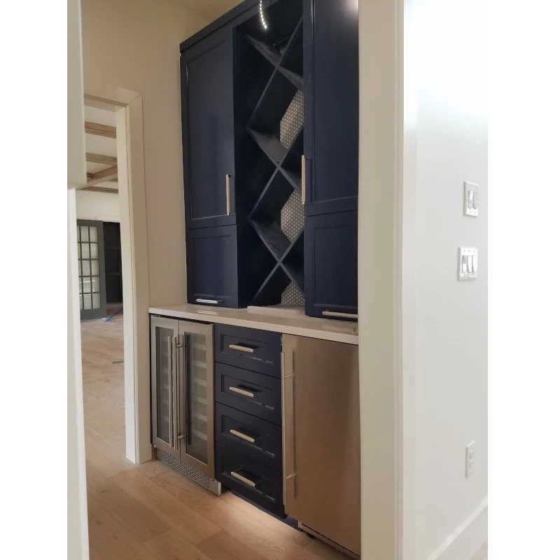 Built-in bar cabinet with dark blue upper cabinets, open wine rack, and lower storage with drawers, a small wine fridge, and a mini fridge, in a kitchen or dining area.