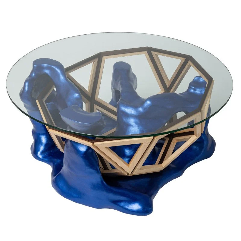 A modern coffee table with a glass top, a geometric wooden frame, and a blue sculptural base with organic shapes.