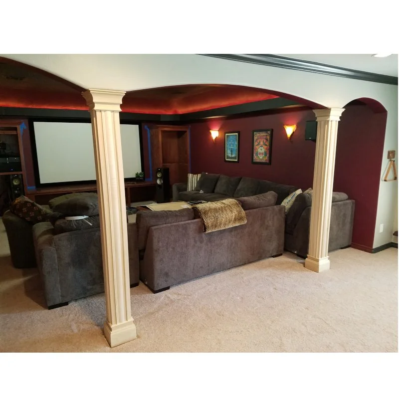 A home theater room with a large sectional sofa, a projection screen, and framed art on a burgundy wall, divided by decorative columns.