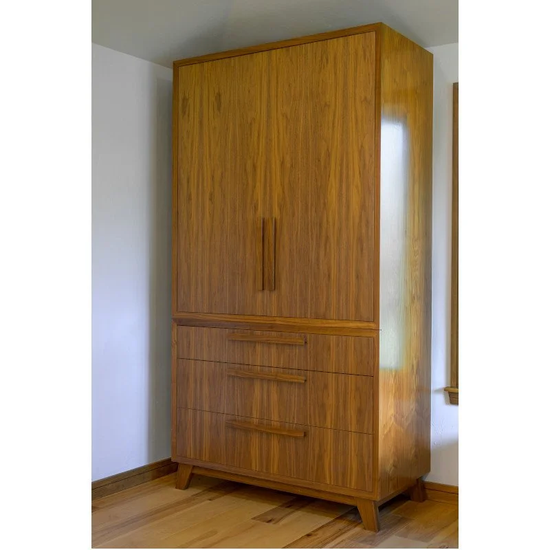 A tall wooden appliance cabinet with three drawers at the bottom and two doors above, featuring a natural wood finish and simple vertical handles, placed against a white wall and standing on a hardwood floor.