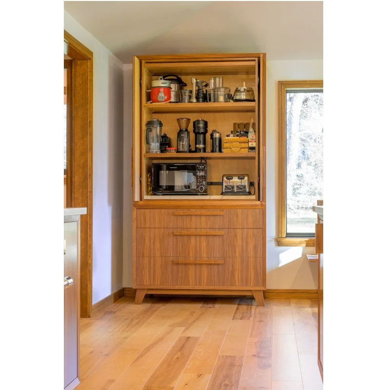 A wooden kitchen cabinet with open shelves on top and drawers below, containing various kitchen appliances and utensils, situated next to a window with natural light.