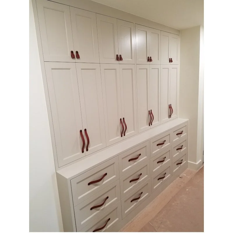 White built-in storage cabinet with multiple upper and lower drawers, featuring brown leather strap handles in a room with light-colored walls and a wooden floor.