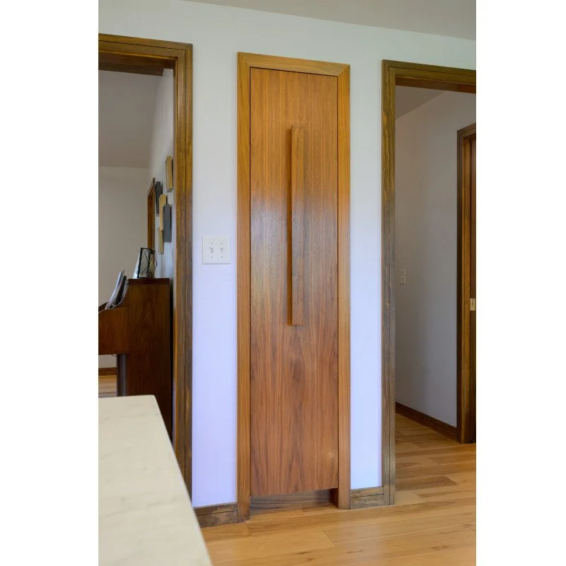 A recessed wooden pantry cabinet with a long vertical handle, located between two doorways in a home interior.