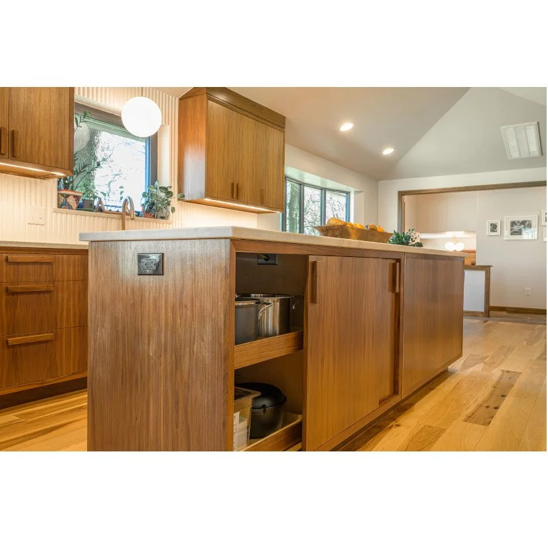 A kitchen with wooden cabinets and a white countertop. There are windows letting in natural light, and the floor is also made of wood.