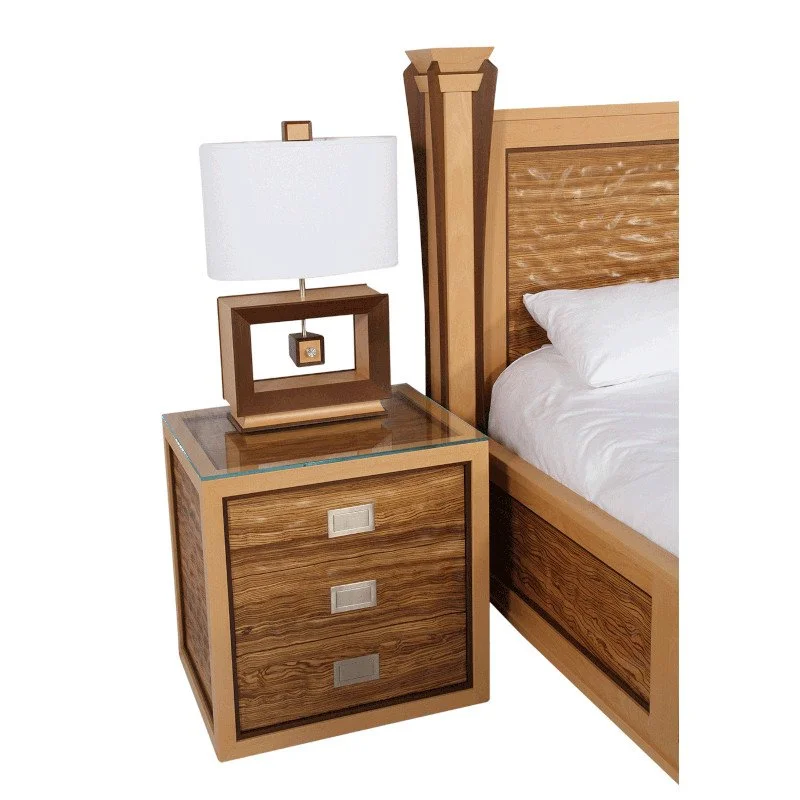 Wooden nightstand with three drawers, a modern table lamp with a white shade, and part of a wooden bed with a headboard and white bedding.