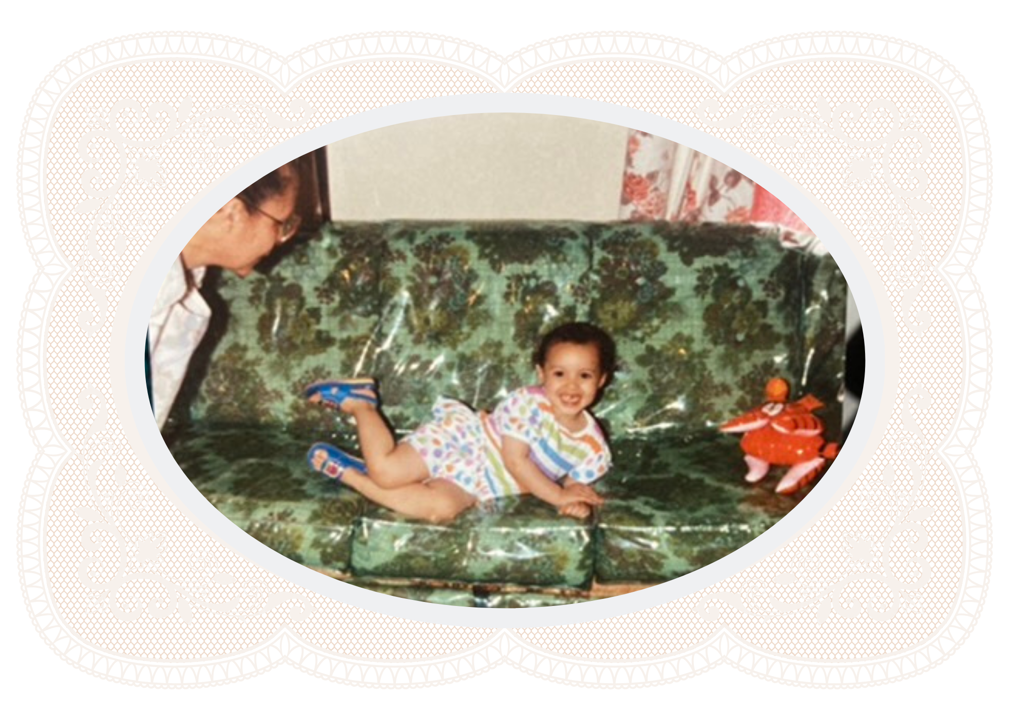 Smiling child lying on a floral-patterned couch beside a red stuffed toy, with an adult partially visible.