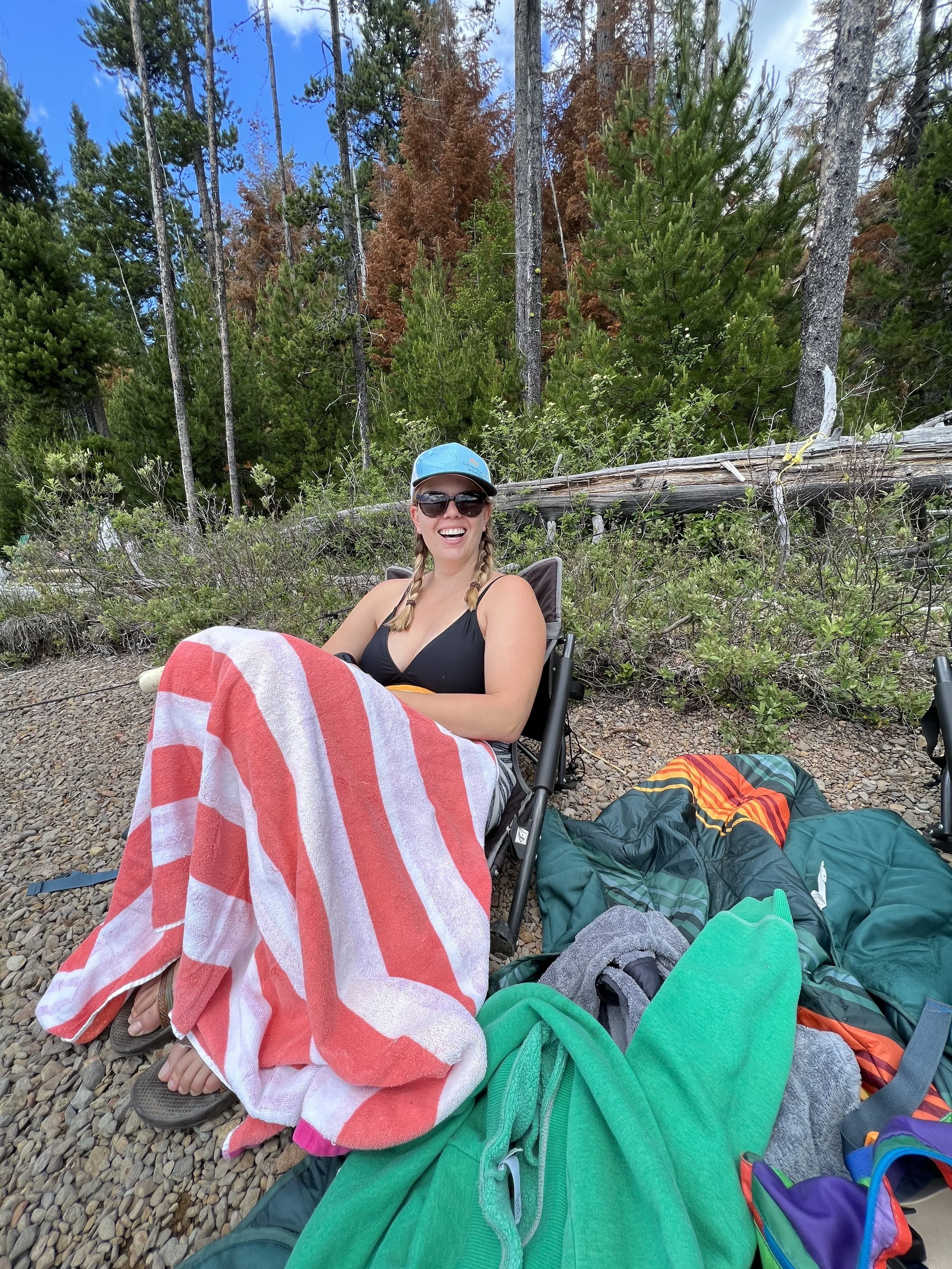 A woman with blonde hair in braids, wearing sunglasses, a blue cap, and a black tank top, smiling while sitting on a camping chair outdoors in a forested area during daytime. She is surrounded by colorful clothing and gear, with tall evergreen trees and blue sky in the background.