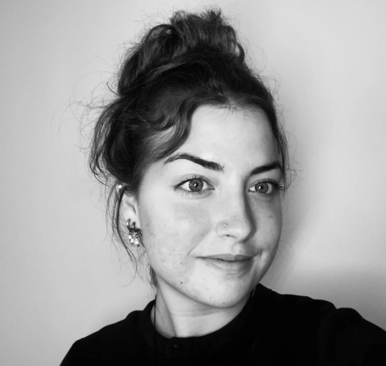 Black and white portrait of a young woman with hair in a messy bun, wearing earrings and a nose piercing, against a plain background.
