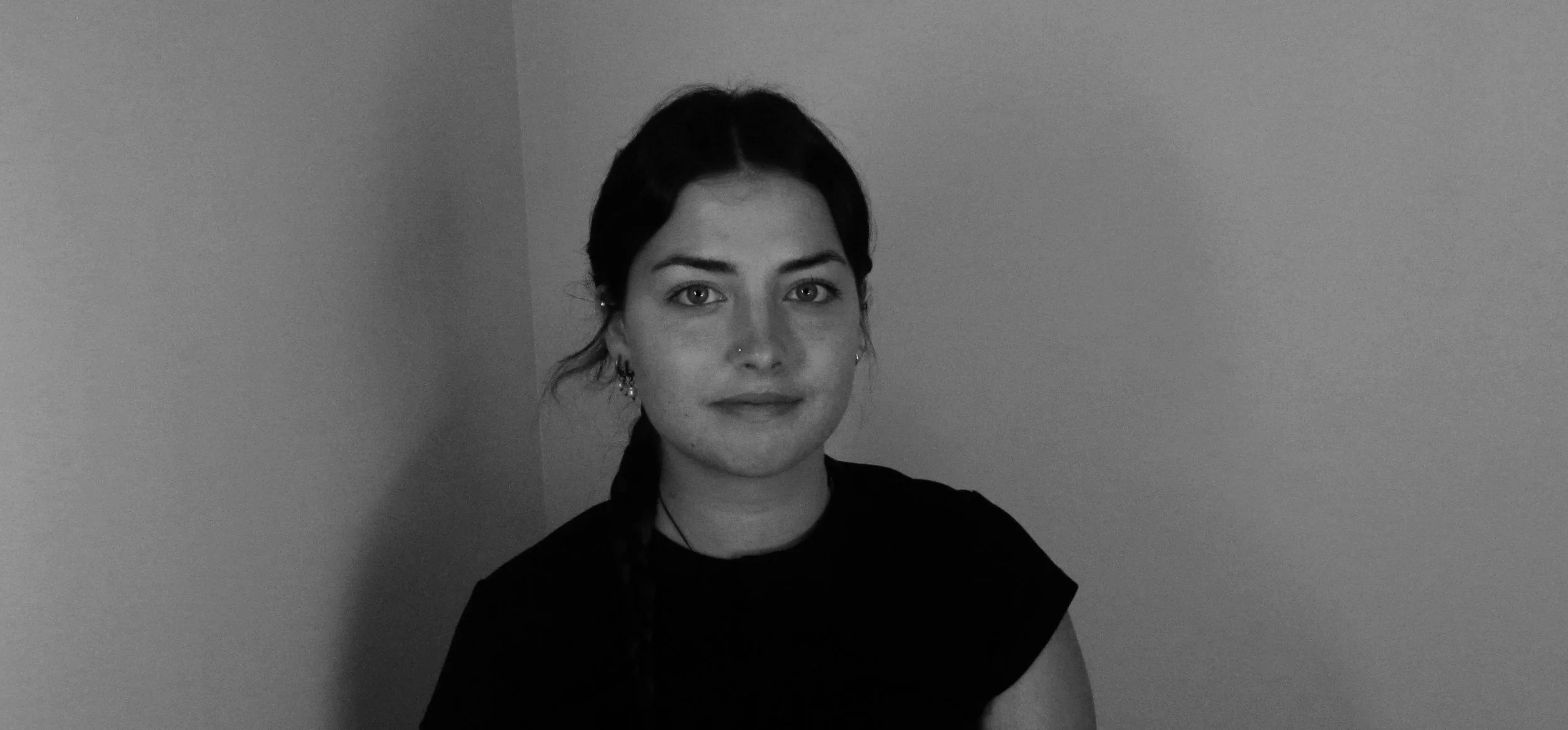 Black and white photo of a young woman with dark hair, wearing a nose ring and earrings, sitting against a plain wall.