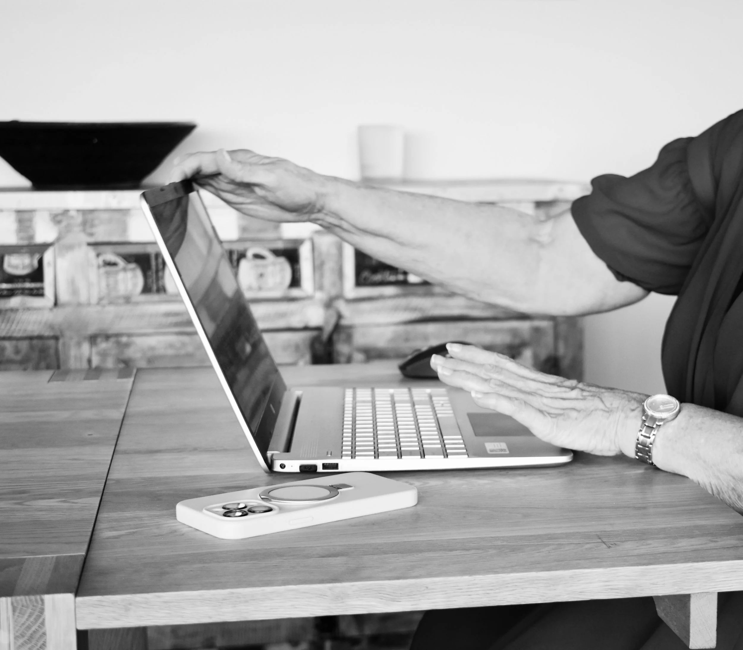 A person with a watch on their left wrist is using a laptop on a wooden table, with one hand on the keyboard and the other on the laptop's top cover. A smartphone with a pop socket is placed on the table nearby.