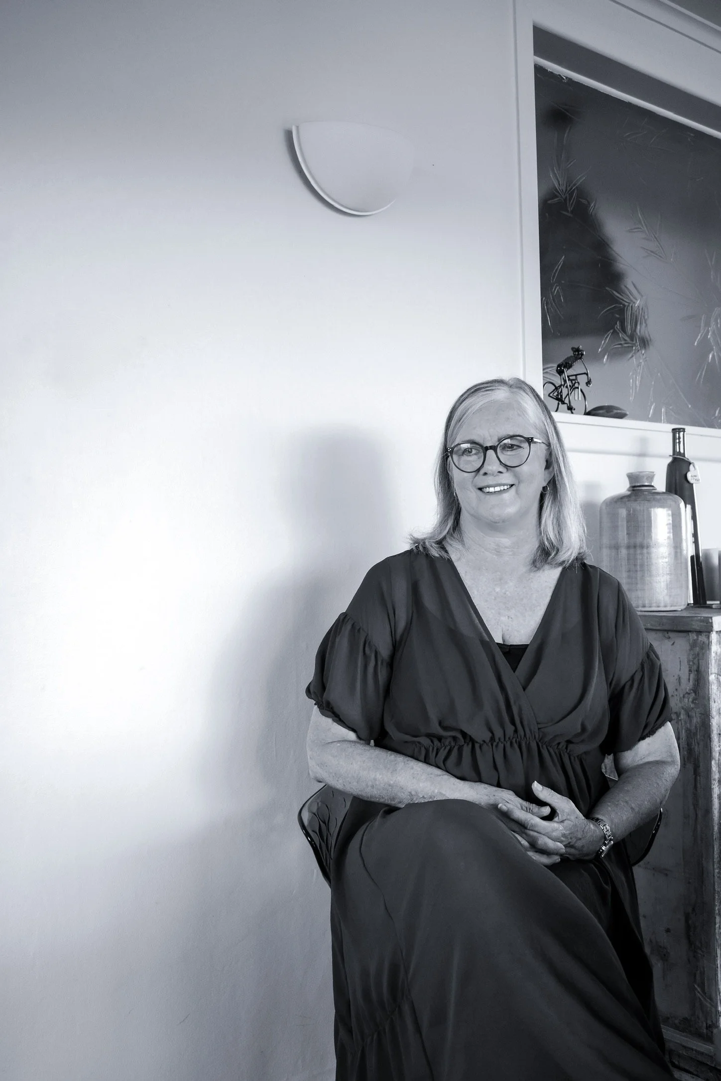 A woman with glasses and shoulder-length hair sitting indoors on a chair, wearing a dark dress, with a wall and some decorative items in the background.