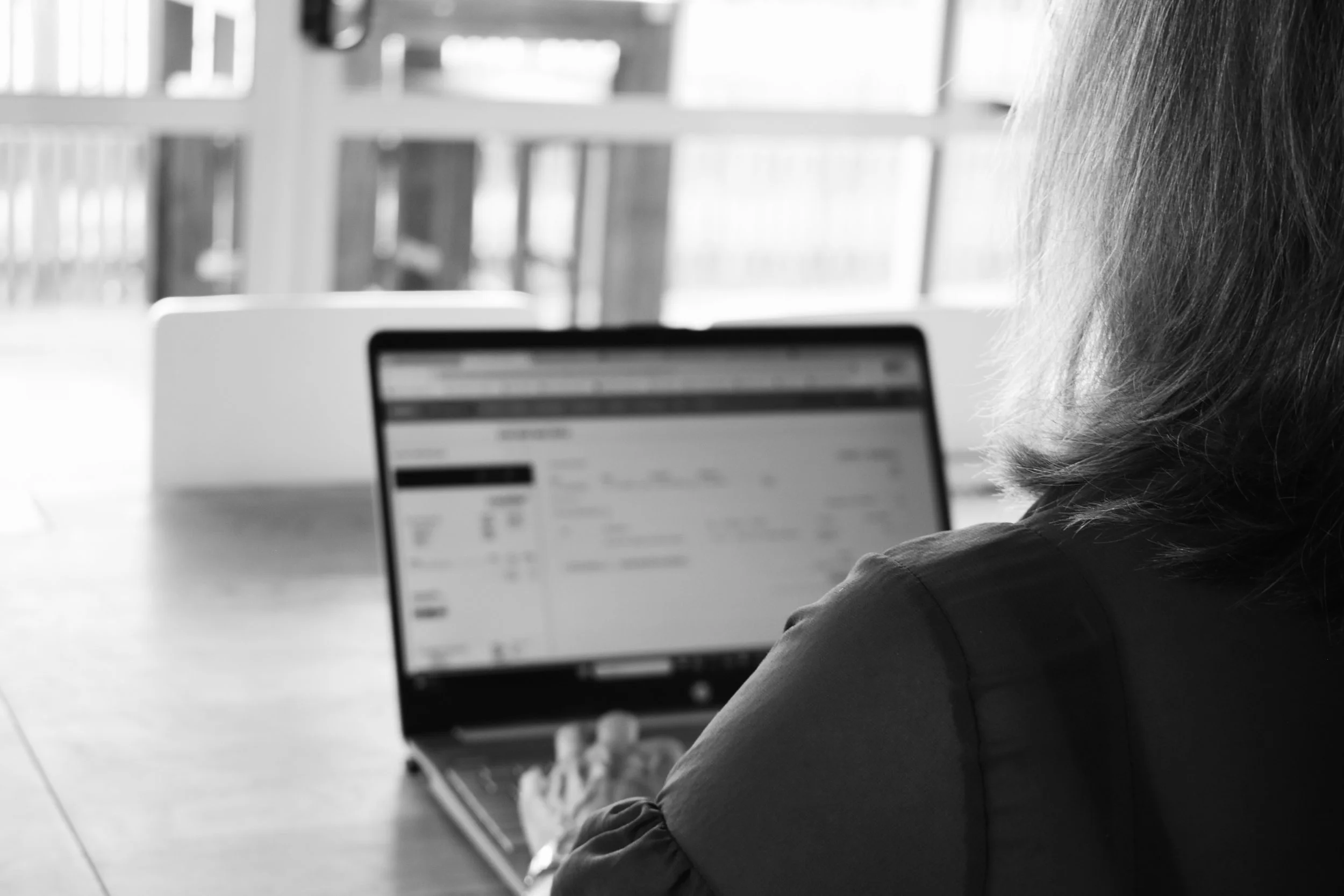 A person working on a laptop at a wooden table near large windows with a view outside.