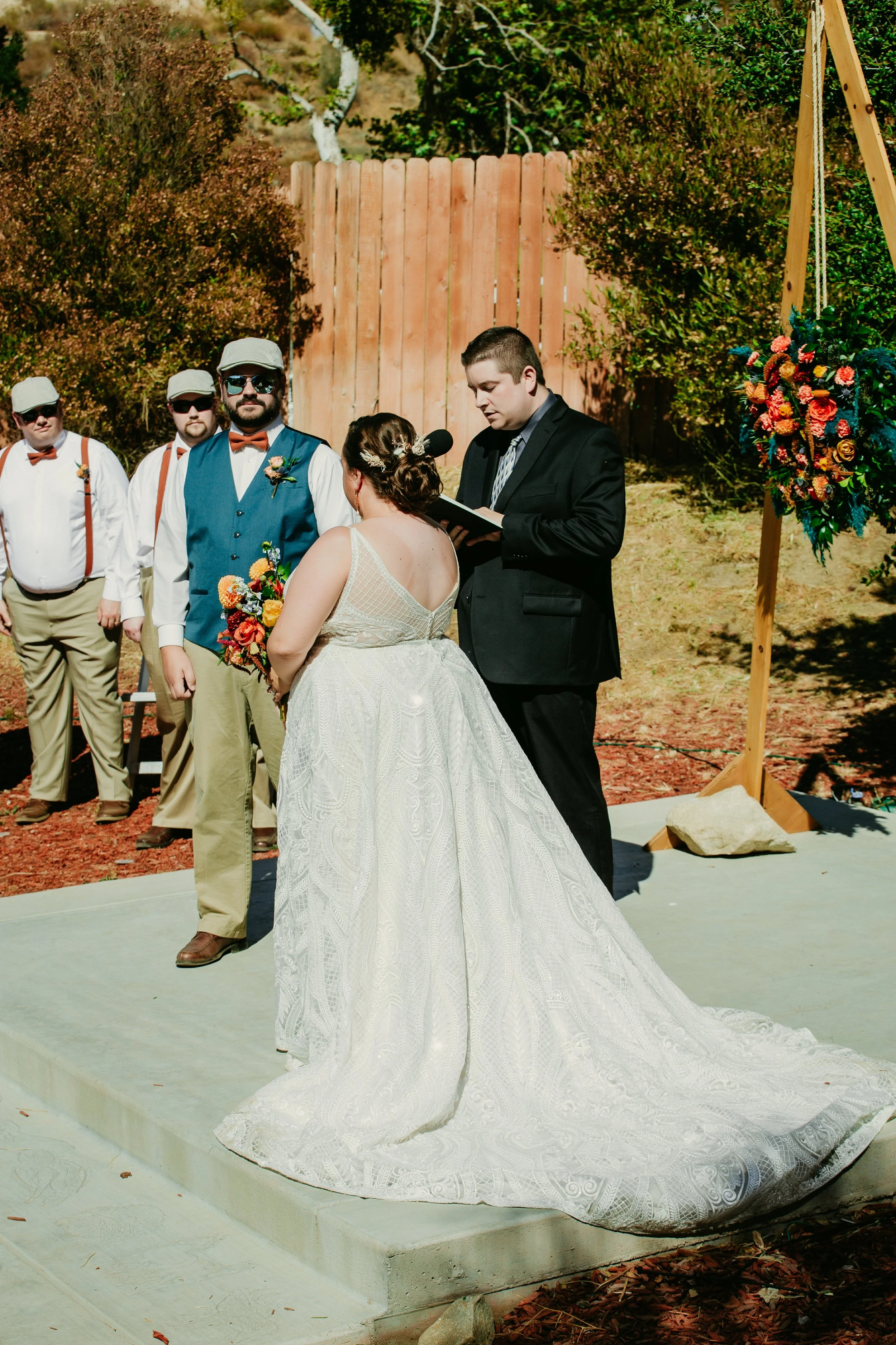 A couple getting married outdoors, facing each other with an officiant reading from a book.
The bride wears a detailed white wedding gown with a train, and the groom in a black suit.
Three groomsmen stand in the background dressed in white shirts, beige pants, orange bow ties, and suspenders.
There are trees, bushes, and a wooden fence in the background, with a colorful floral arrangement on a wooden stand to the side.