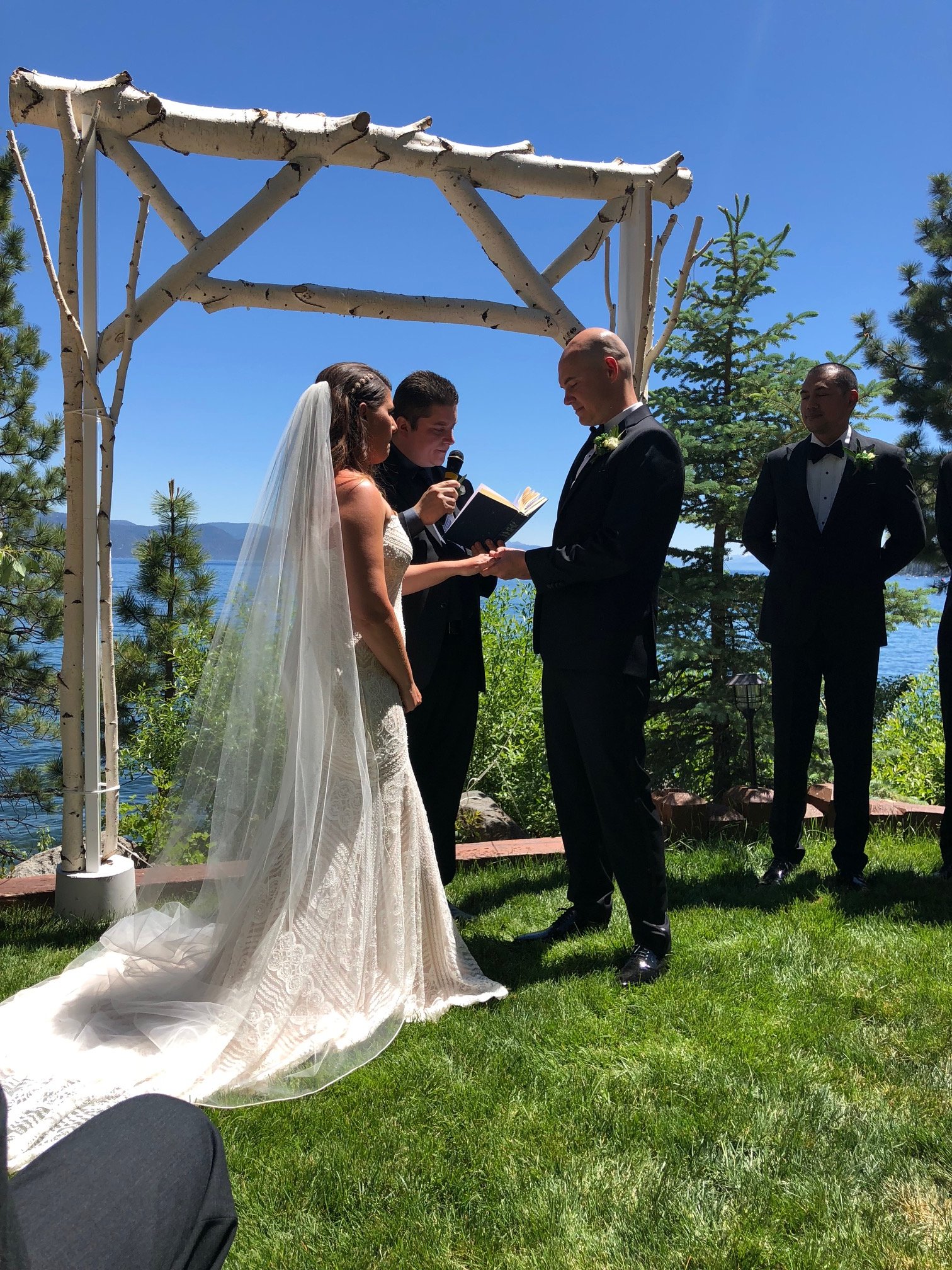 A wedding ceremony outdoors near a lake with a blue sky, where a bride and groom are exchanging vows under a wooden arch with officiant reading from a book, surrounded by groomsmen and pine trees.