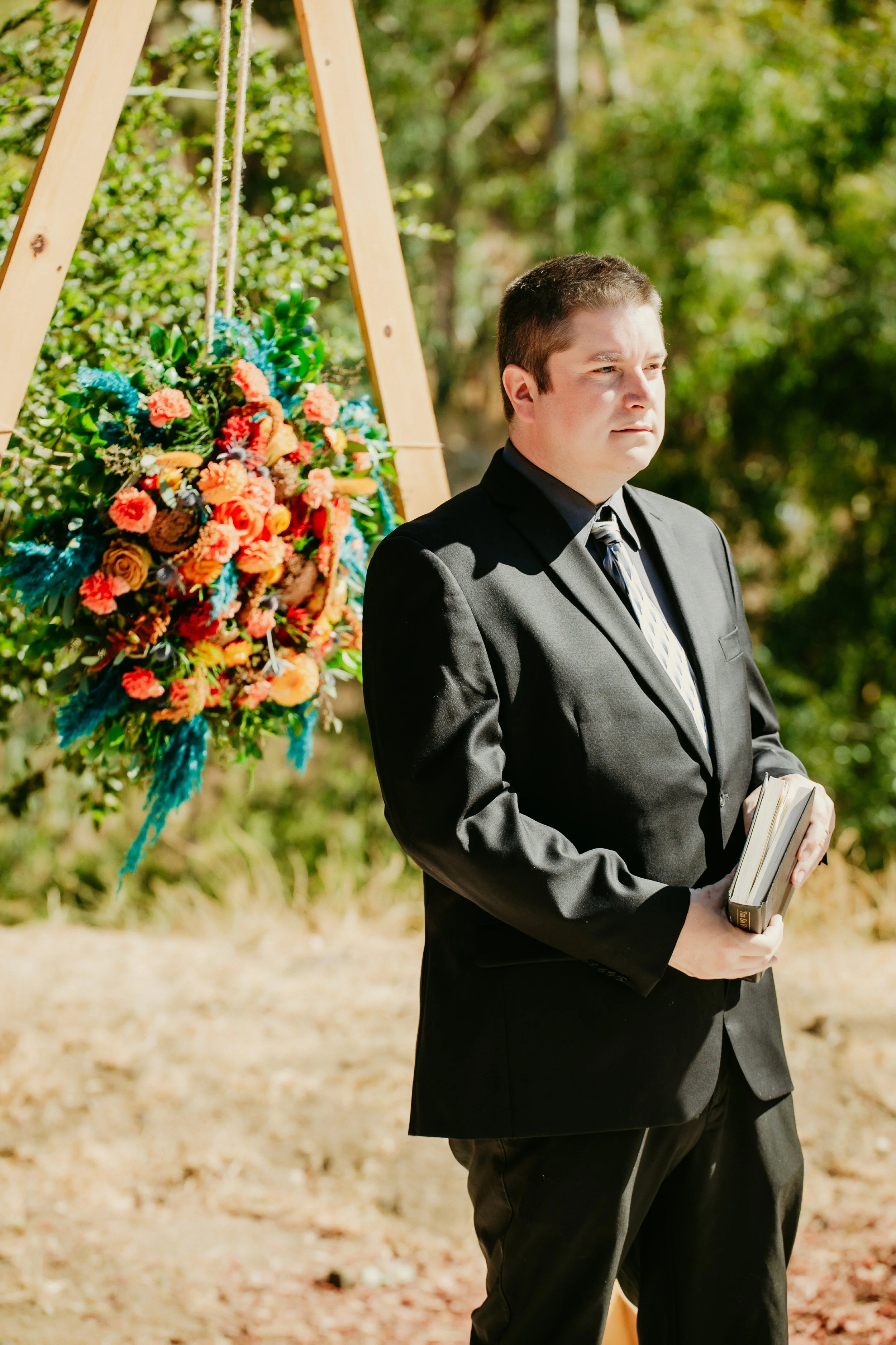 Reverend James in a black suit and tie standing outdoors holding a book during a ceremony, with a colorful floral arrangement on a wooden stand behind him.
