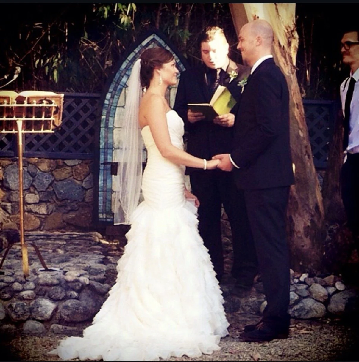 A wedding ceremony outdoors where a bride and groom are exchanging vows under a wooden arch with officiant reading from a book, surrounded by groomsmen.