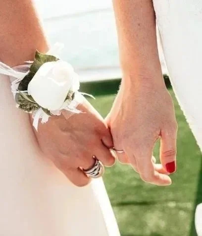 Two people holding hands, one wearing a wedding ring and a white rose boutonniere.