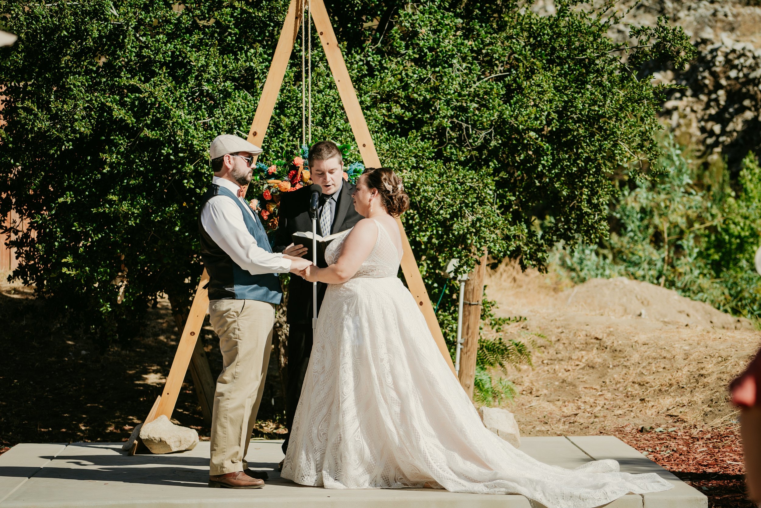 A couple getting married during an outdoor ceremony, with the officiant standing between them. The bride is in a white dress, and the groom is in a blue suit. The setting appears to be a garden or park with trees and greenery in the background.