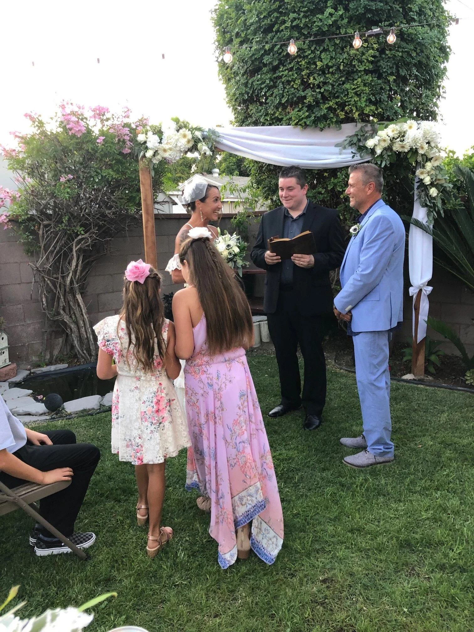 A backyard wedding ceremony with a bride and groom standing under a decorated arch with white flowers, two young girls in floral dresses with pink and white bows, and a man in a light blue suit officiating. The setting is outdoors with greenery and string lights overhead.